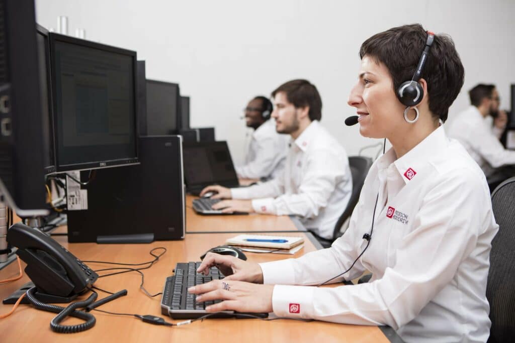 Female wearing a phone headset and sat in front of a desktop computer. In the background, other team members are sat at desks working.