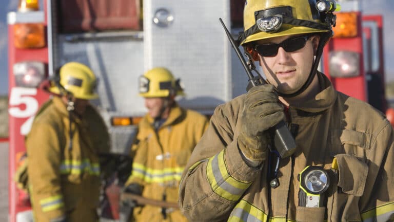 Firefighter talking on radio with colleagues standing by at the back of a fire engine in the background