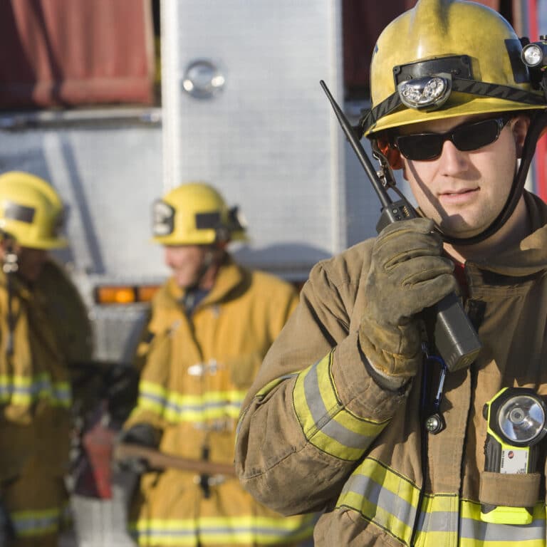 Firefighter talking on radio with colleagues standing by at the back of a fire engine in the background