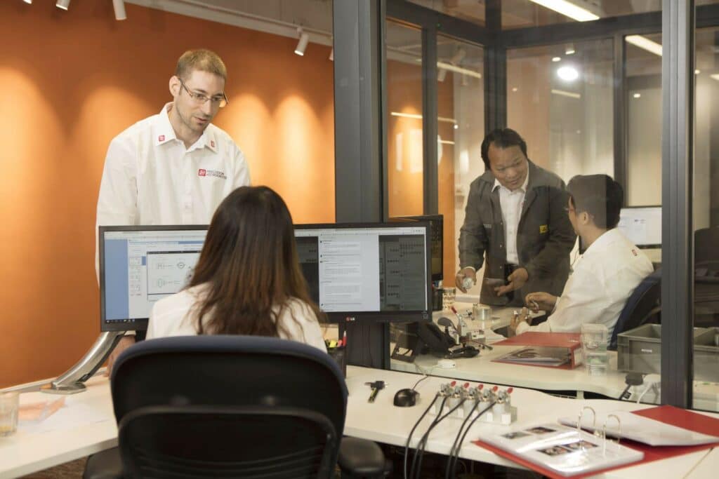 A PMD team member sat in front of a computer (but with their back the to camera) discussing a project with a colleague. To their right two more colleagues are discussing motor specifications. 