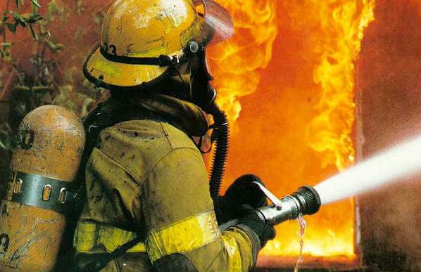  The side view of a firefighter dressed in all their protective equipment holding a hose with a burning fire in the background.