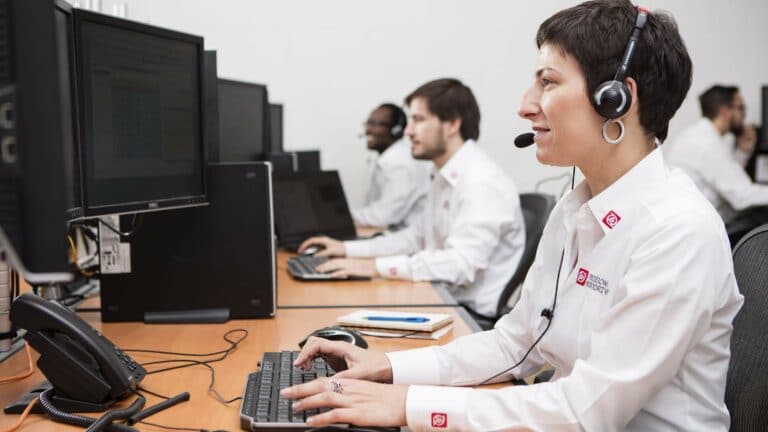 Woman sat in from of a computer with a headset on with three of her colleagues working at computers in the background