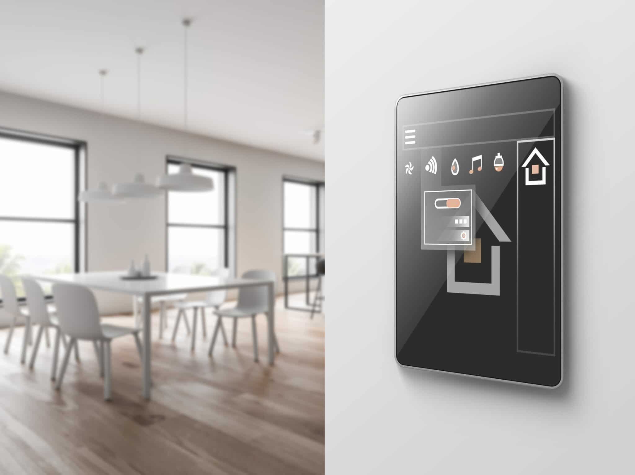 In the foreground there is a touchscreen control panel for smart blinds mounted to a wall. In the background is a dinning room with blinds installed in the windows.